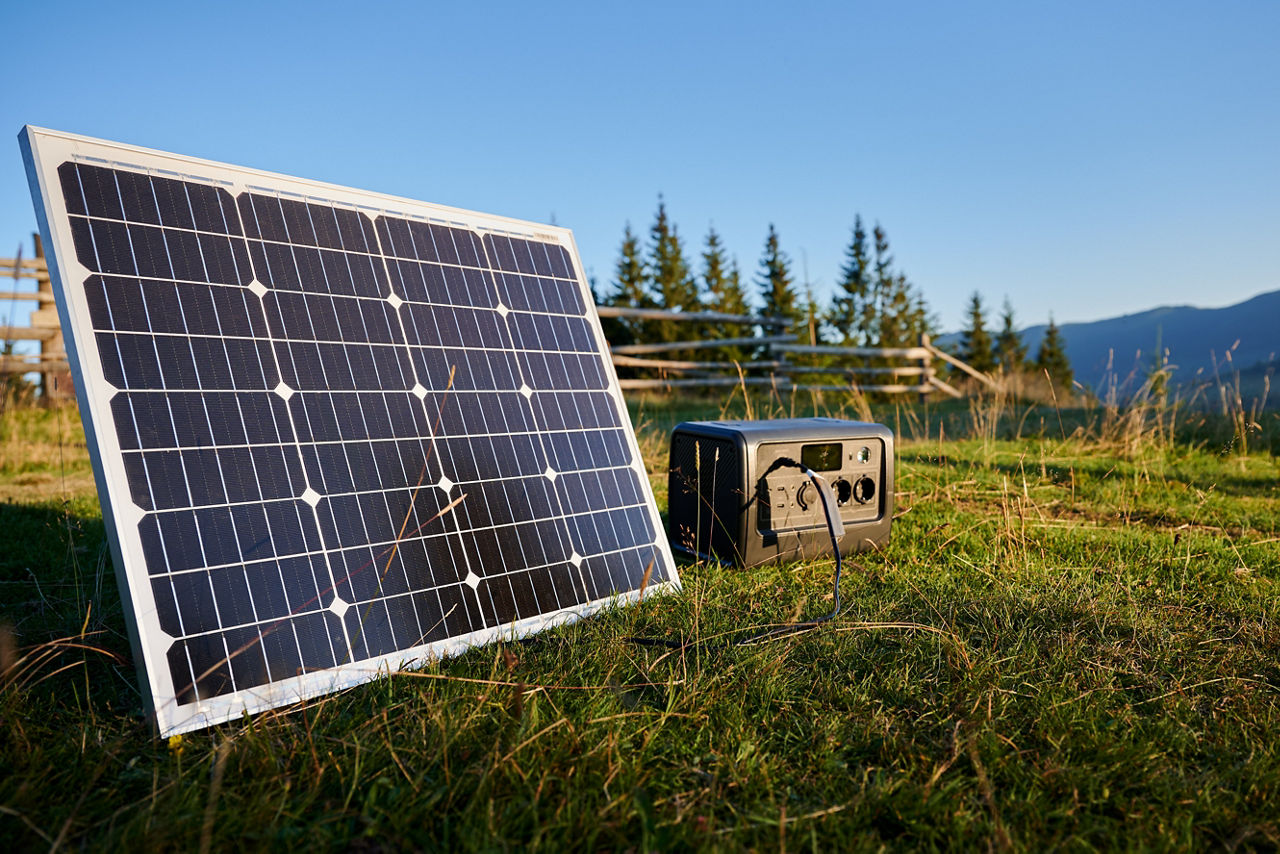Tourist tent with solar panel and portable power station nearby in grassy field. Scene surrounded by rolling hills and wooden fence under clear blue sky, showcasing eco-friendly camping setup.