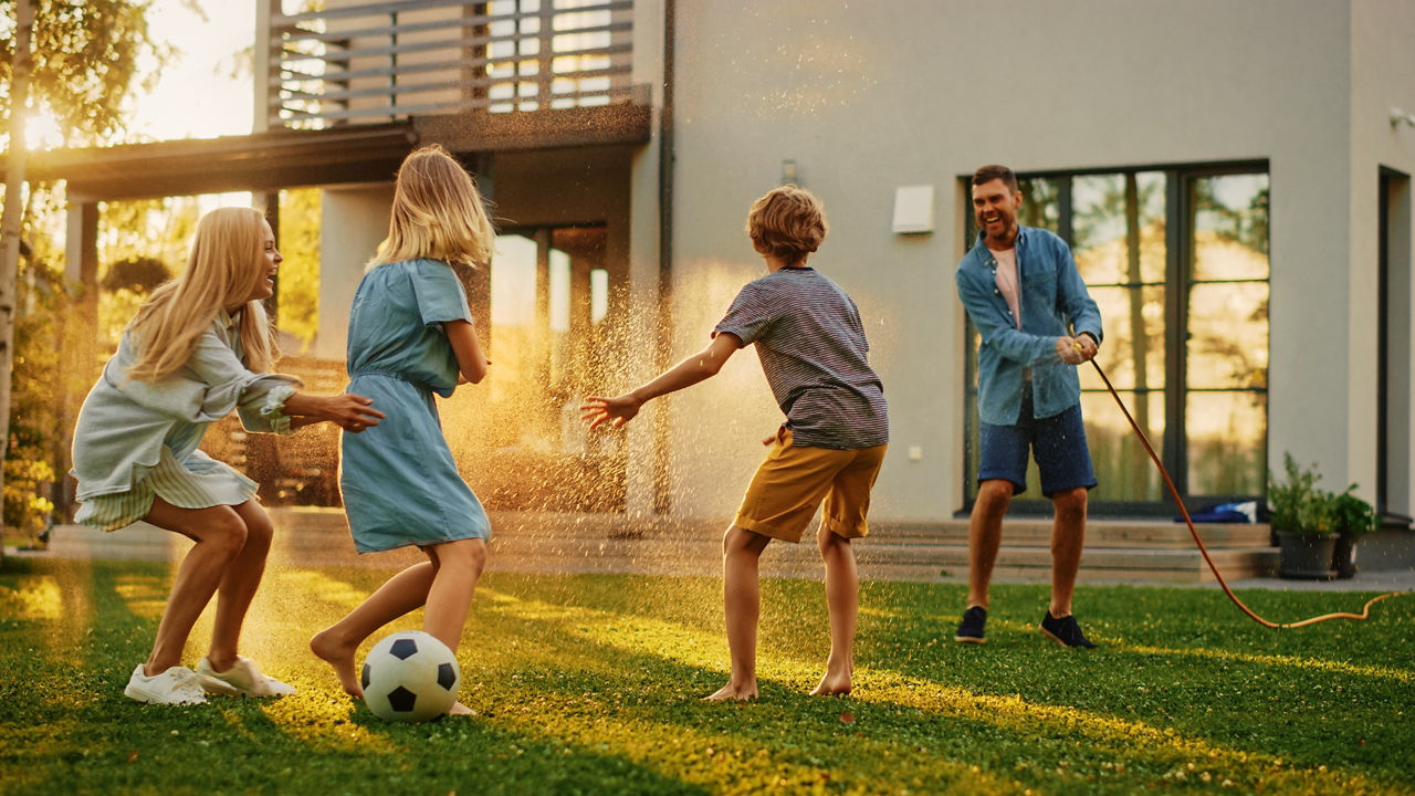 Glückliche vierköpfige Familie, die mit Gartenwasserschlauch spielt und sich gegenseitig bespritzt. Mutter, Vater, Tochter und Sohn haben Spaß beim Spielen auf dem Hinterhofrasen eines idyllischen Vorstadthauses an einem sonnigen Sommertag