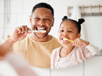Child, dad and brushing teeth in a family home bathroom for dental health and wellness in a mirror. Face of african man and girl kid learning to clean mouth with toothbrush and smile for oral hygiene.