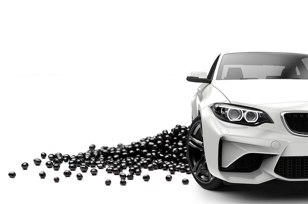 Black shiny polymer pellets are laid on white background, in front of it, a white sports car is placed 