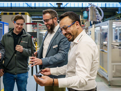 Happy employees interacting with product sample, holding EV coolant hose in. hands, employees starting left is in a grey sweater with a Teknor Apex emblem: Tim Neckermann; in the middle in a light gray suit with a name tag: Marco Eulenbach; on the forefront with a white button down t-shirt and Teknor Apex logo stiched onto the collar: Ali Kaya